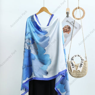 A floral design satin silk scarf in blue and white, displayed on a wooden hanger, with a casual outfit in the background.
